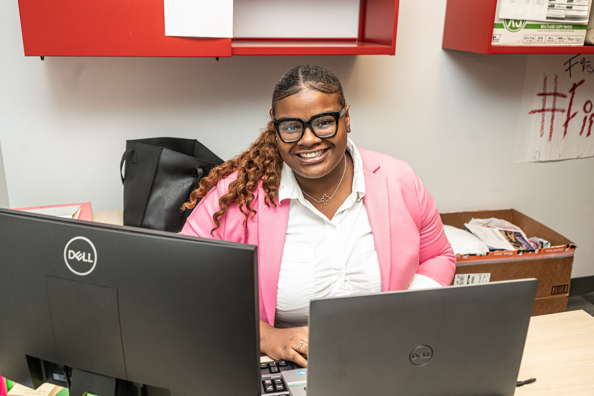 Peron wearing a button-up blouse and pink jacket working on a laptop with an extra monitor.