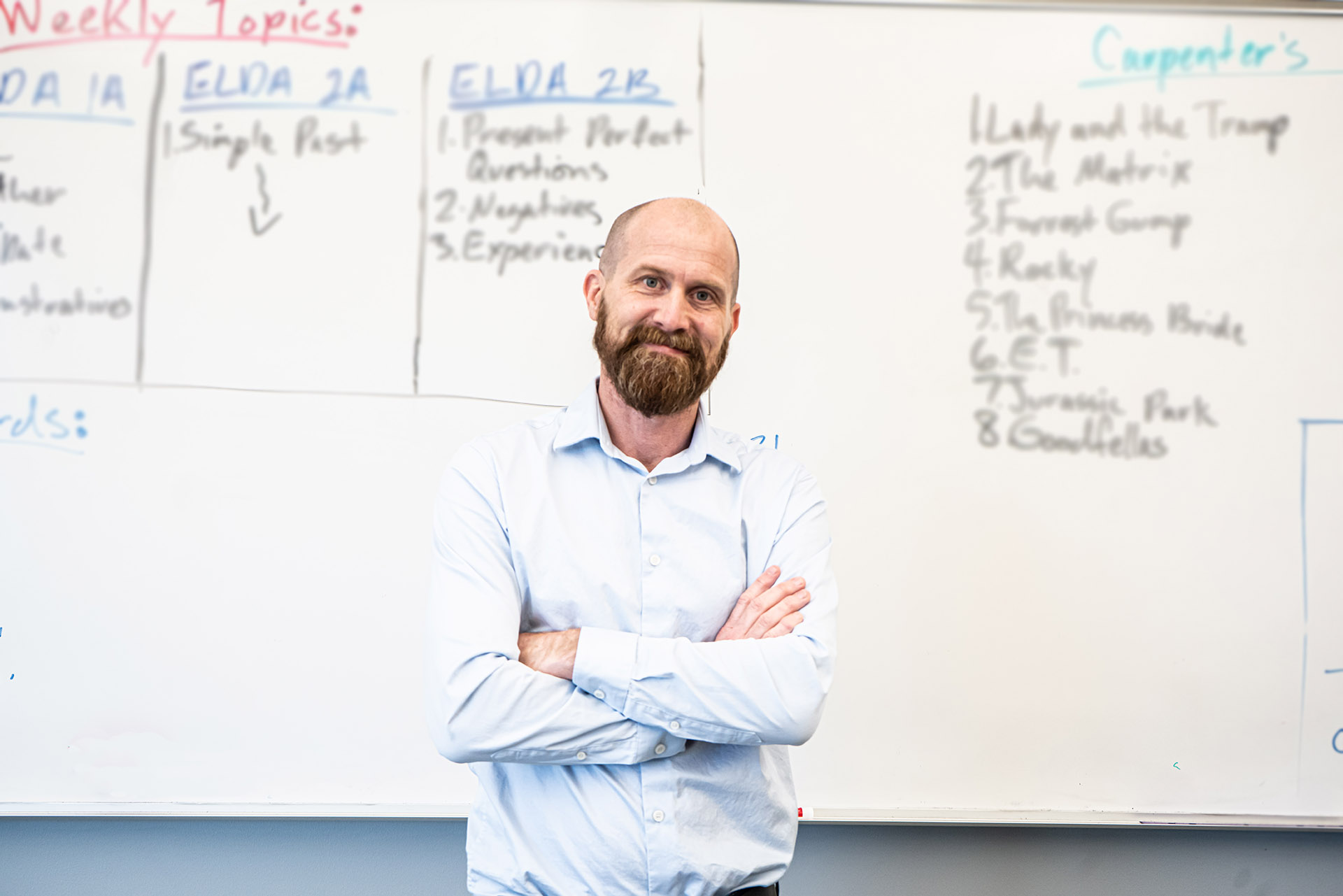 Person with a beard wearing a button up shirt and smiling in front of a whiteboard.