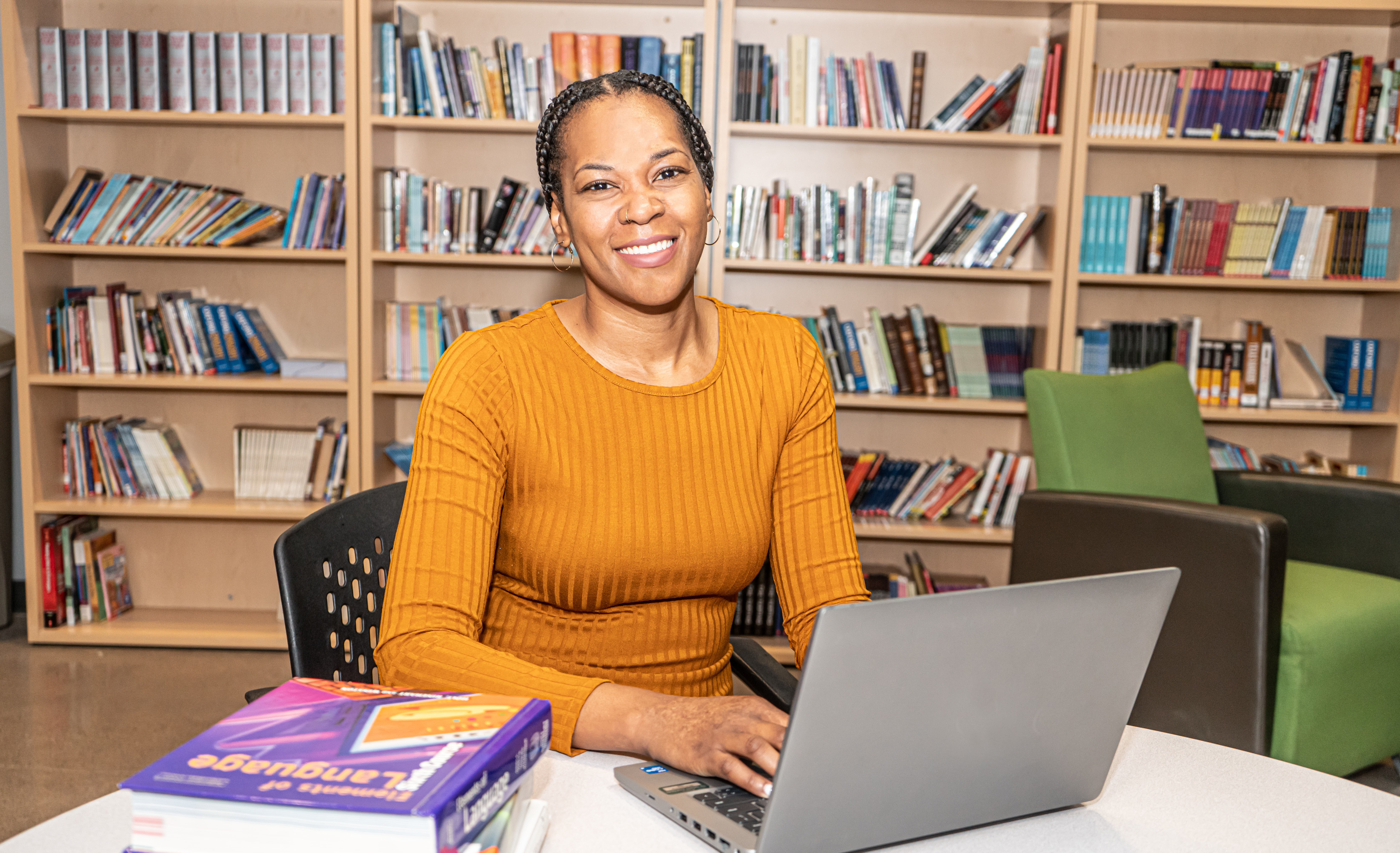 Person in a fitted sweater sitting at a library table working on a laptop and smiling.