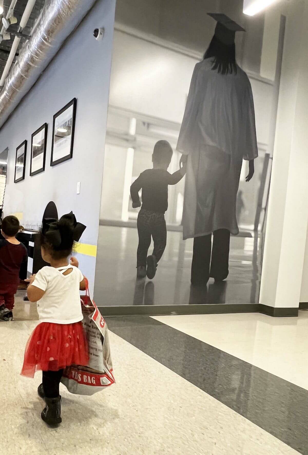 Toddler walking down the Excel School hallway beside a mural of a graduate with their child.